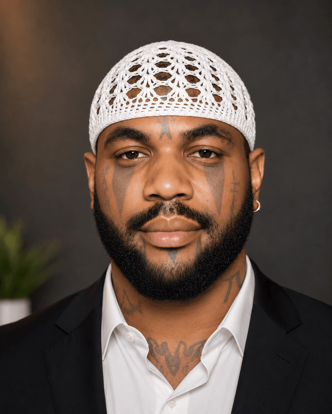 Bearded Black man with facial tattoos wearing a white crochet skullcap and black suit.
